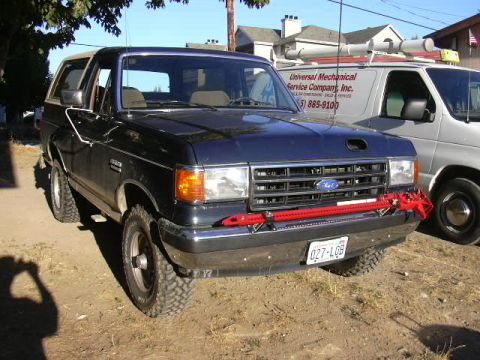 1991 Ford Bronco - Front three-quarter view of a late-1980s Ford Bronco in dark paint with a tan removable hardtop, a red Hi-Lift style jack mounted across th…