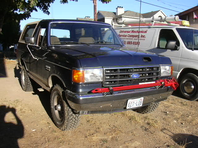 Dark late-80s Ford Bronco with tan top and red jack on front bumper, parked by a work van