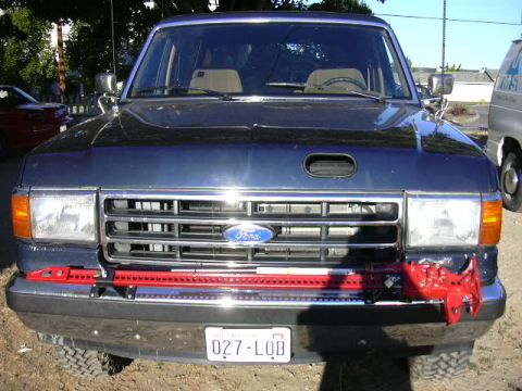 1991 Ford Bronco - Front view of a dark blue 1980s-90s Ford SUV with chrome grille bearing the Ford oval, a red front tow/mount bracket across the bumper, and…