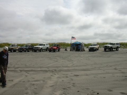 1991 Ford Bronco - Several vehicles are parked on a sandy beach with dunes and a cloudy sky overhead.