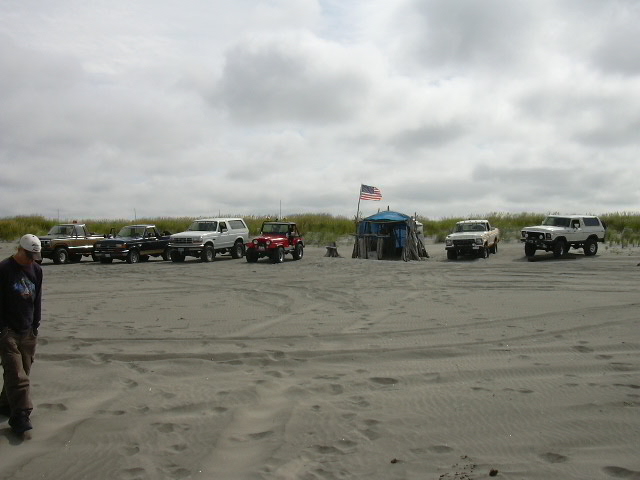 Row of 4x4 trucks and a Jeep parked on a beach around a blue canopy with a US flag