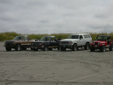 1991 Ford Bronco - A white 1991 Ford Bronco is parked on the sand alongside three other off-road vehicles, with dunes and a cloudy sky in the background.