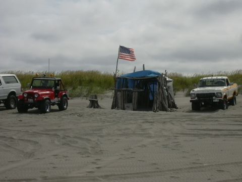1991 Ford Bronco - Two off-road vehicles and a makeshift shelter on a sandy beach under a cloudy sky.