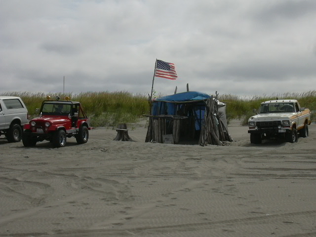 Ford pickup truck and red Jeep parked on sand near a driftwood shelter with American flag