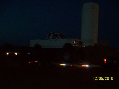 1985 Ford Bronco - A white pickup truck is loaded onto a trailer at night, with a silo visible in the distance.