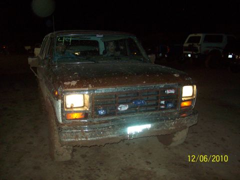 1985 Ford Bronco - Muddy 1980s Ford truck with headlights on, seen at night with another vehicle in the background.