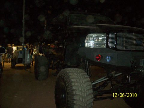 1985 Ford Bronco - A lifted black truck is seen at night with large tires and an aftermarket bumper, with other vehicles and people in the background.