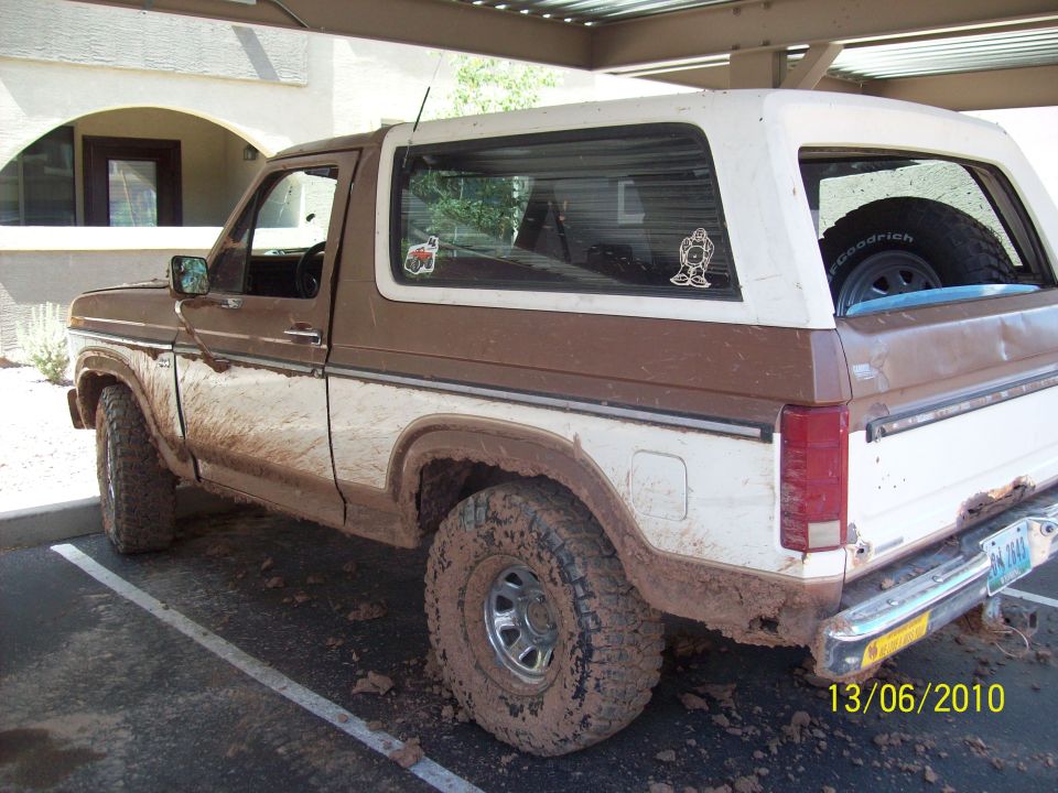 Mud-covered 1980s Ford Bronco parked under a carport, showing its side profile and rear