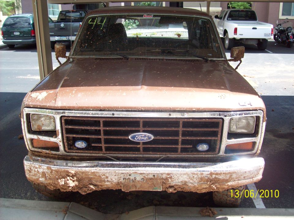Brown Ford truck with a rusty grille and bumper covered in mud. A white pickup and a motorcycle are parked behind it