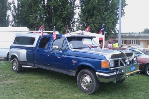 1991 Ford Crown Victoria - A blue and silver dually pickup truck with a camper shell and decorative flags is parked on a grassy field.