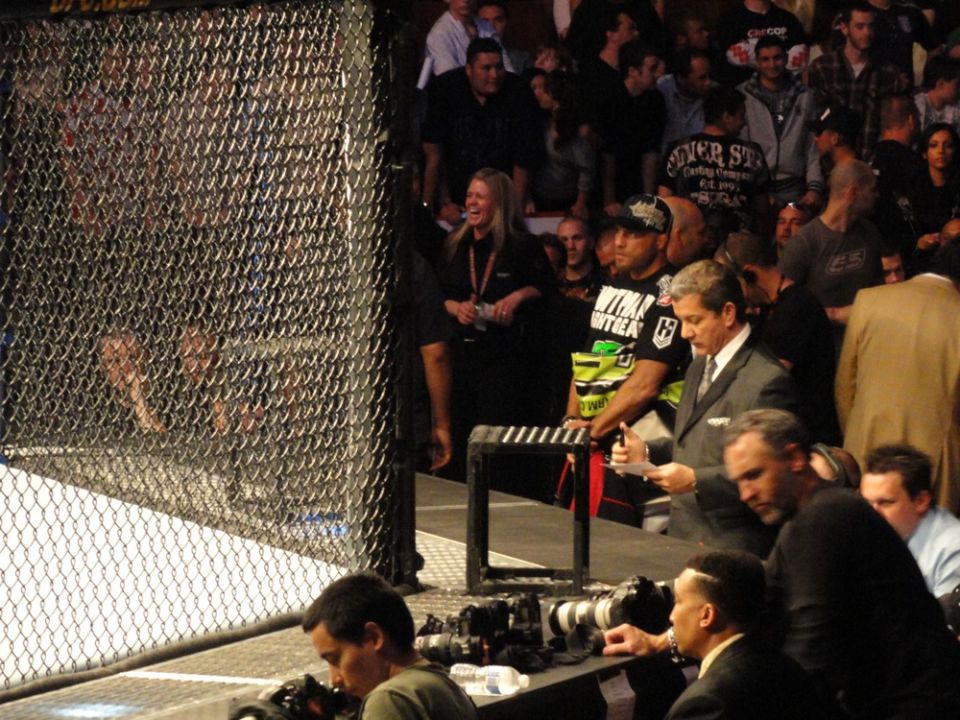 Spectators behind a chain-link fence at an indoor event, with some people in suits and others in casual attire