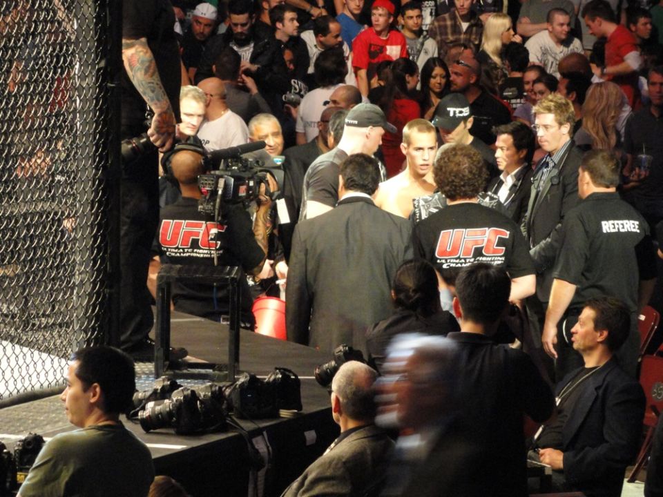UFC fighters in ring, surrounded by officials and crowd. Camera crew filming