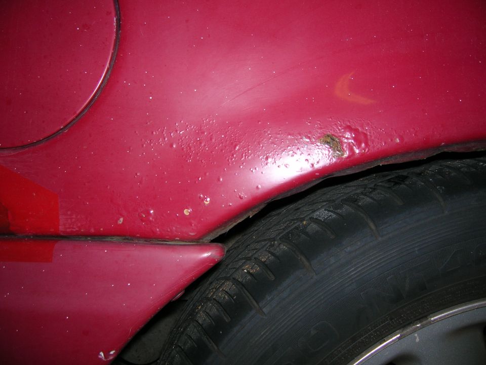 Close-up of a red car's rear fender showing bubbling paint and rust near the wheel well