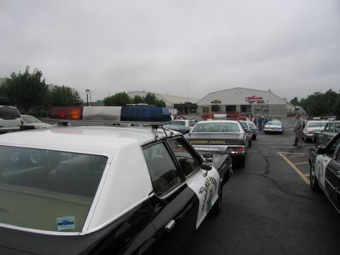1991 Ford LTD Crown Victoria - black-and-white police cruiser in the foreground among a lineup of patrol cars in a gray, overcast parking lot near warehouses.