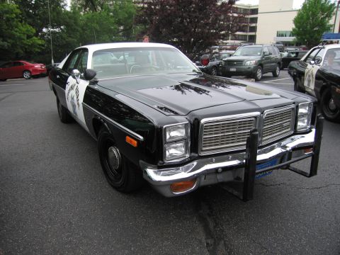 1991 Ford LTD Crown Victoria - black 1970s-style police sedan with chrome grille, push bumper and a door decal, parked in a lot with other vehicles behind.