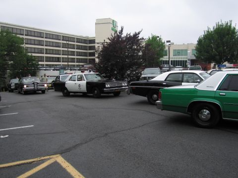 1991 Ford LTD Crown Victoria - black-and-white police sedan moving through a parking lot, with a green-and-white cruiser in the foreground and an office building behind.