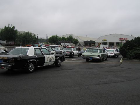 1991 Ford LTD Crown Victoria - black-and-white highway patrol sedan beside a green-and-white station wagon in a gray overcast lot with people gathered behind.