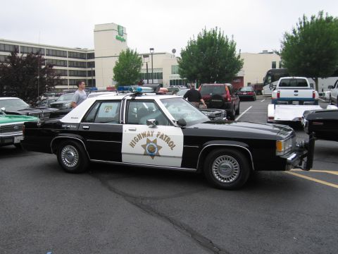 1991 Ford LTD Crown Victoria - black-and-white highway patrol Ford sedan with door lettering and roof light bar, parked in a lot with trucks and a building behind.