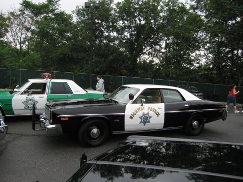 1991 Ford LTD Crown Victoria - black-and-white highway patrol sedan parked next to a green-and-white cruiser along a chain-link fence, with people walking by.