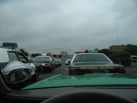 1991 Ford LTD Crown Victoria - Dashboard view through a windshield showing black highway patrol sedans with light bars parked in a lot under an overcast sky.