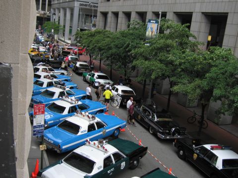 1991 Ford LTD Crown Victoria - Elevated view of a city street lined with blue-and-white NYPD cruisers and other vintage police cars at a display, with crowds and trees.