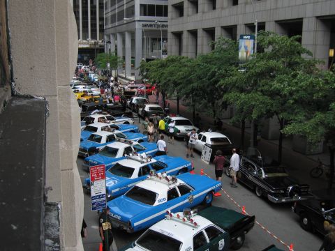 1991 Ford LTD Crown Victoria - Overhead view of blue-and-white NYPD sedans and assorted police cars parked along a tree-lined downtown street with onlookers.
