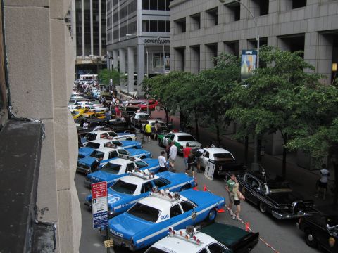 1991 Ford LTD Crown Victoria - Elevated view of a downtown street lined with blue-and-white NYPD sedans, a yellow taxi and other police cars amid a crowd.