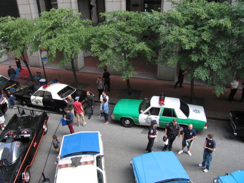 1991 Ford LTD Crown Victoria - Elevated street view of a green-and-white police wagon and black cruisers with officers and a crowd gathered under trees.