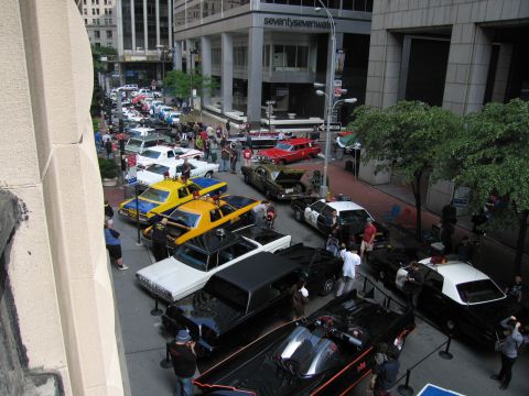 1991 Ford LTD Crown Victoria - Elevated view of a downtown street display of vintage vehicles, including yellow taxis, a black car and onlookers among the buildings.