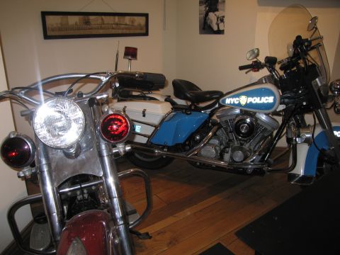 1991 Ford LTD Crown Victoria - Two vintage police motorcycles displayed indoors on a wood floor, the rear one in blue with NYC POLICE lettering on the tank.