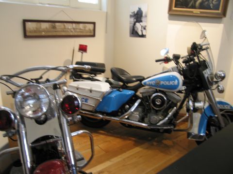 1991 Ford LTD Crown Victoria - blue-and-white NYC Police touring motorcycle with a windshield and saddlebags sits on a wood floor in a museum-style display room.