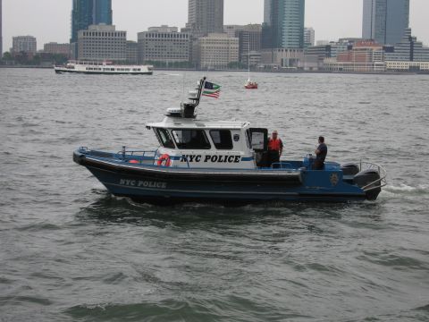 1991 Ford LTD Crown Victoria - blue-and-white NYC Police patrol boat cruises across open harbor water with officers aboard and a city skyline in the background.
