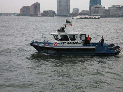 1991 Ford LTD Crown Victoria - NYC Police patrol boat with two officers aboard motors across gray harbor water in front of a distant waterfront skyline.