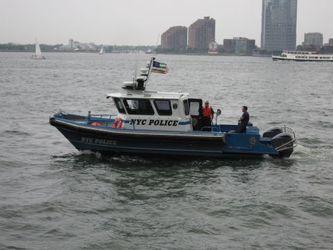 1991 Ford LTD Crown Victoria - blue NYC Police patrol boat with twin outboard motors and crew aboard runs across harbor water near a city waterfront.