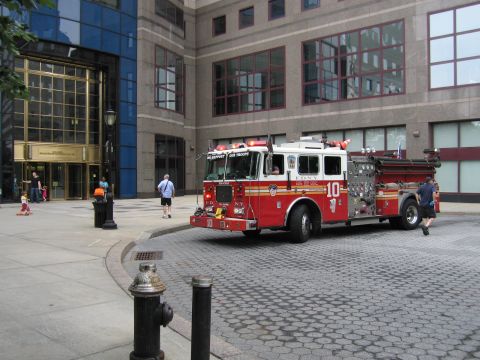 1991 Ford LTD Crown Victoria - red FDNY Engine 10 pumper fire truck is parked at a curb on a cobblestone street beside a tall office building.