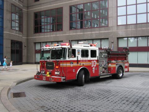 1991 Ford LTD Crown Victoria - red FDNY Engine 10 fire truck marked "We Support Our Troops" sits on a cobblestone street in front of a large glass-and-stone building.