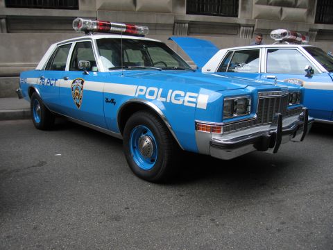 1991 Ford LTD Crown Victoria - vintage blue-and-white POLICE sedan with a chrome push bar and red roof dome lights is parked at an angle beside other cruisers.