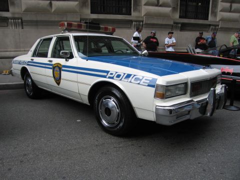 1991 Ford LTD Crown Victoria - white-and-blue Transit Police sedan with a roof light bar and chrome push bar is parked on a city street near onlookers.