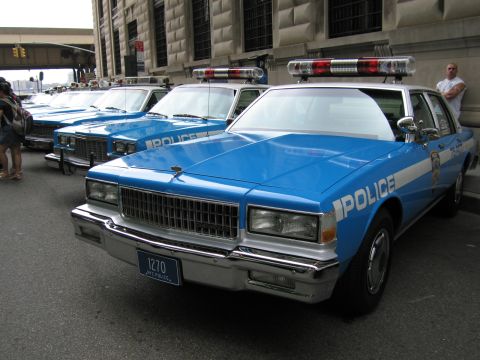 1991 Ford LTD Crown Victoria - blue-and-white NYPD POLICE cruiser with plate 1270 and red roof lights sits at the front of a lined-up row of vintage sedans.