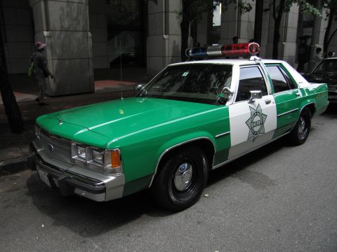 1991 Ford LTD Crown Victoria - green-and-white Sheriff sedan with chrome bumper and roof light bar is parked at a curb with a pedestrian walking behind it.