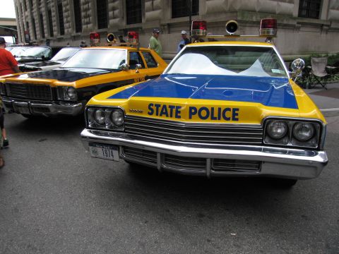 1991 Ford LTD Crown Victoria - yellow-and-blue STATE POLICE sedan with roof lights and a chrome grille is parked at the curb beside a matching cruiser.