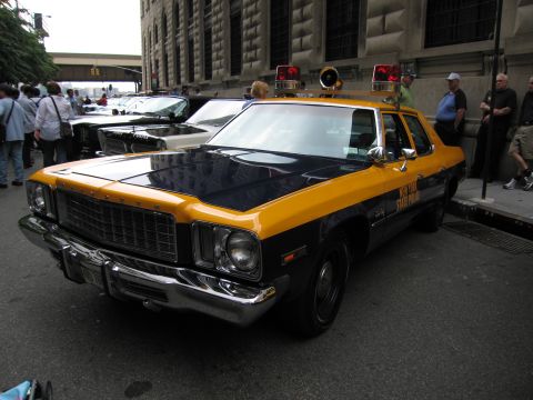 1991 Ford LTD Crown Victoria - yellow-and-black State Police sedan with roof lights sits at the curb amid a crowd and other parked vehicles on a city street.