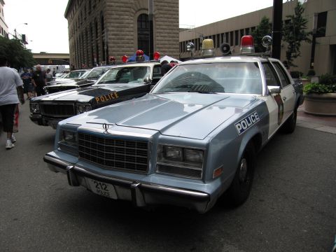 1991 Ford LTD Crown Victoria - silver-and-white POLICE sedan numbered 212 with red roof dome lights is parked at the front of a row of vintage cruisers.