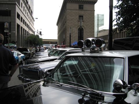1991 Ford LTD Crown Victoria - Close-up of a chrome twin-spotlight and siren cluster mounted on a vintage police car, with a row of cars receding down a city street.