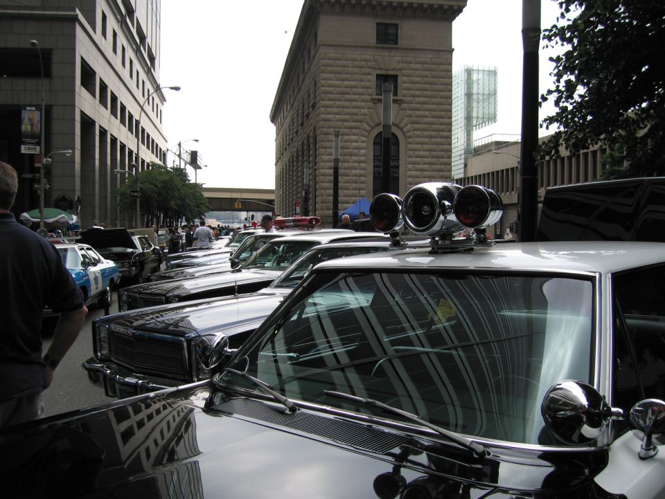 Chrome dual-light police bar and siren atop a classic car at an urban car show