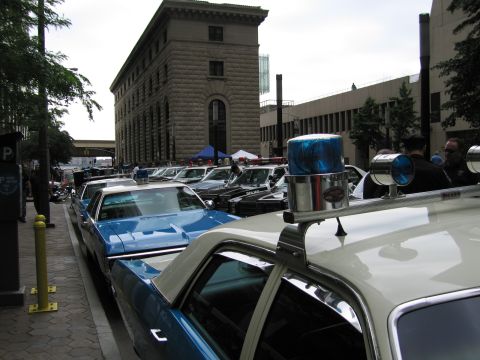 1991 Ford LTD Crown Victoria - long row of classic police sedans, one with a blue roof beacon, parked beside a stone building during a car show.