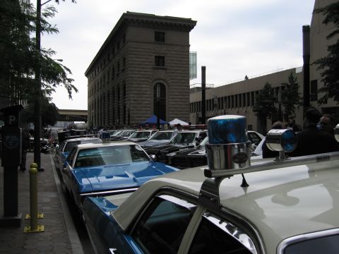 1991 Ford LTD Crown Victoria - Vintage squad cars with chrome and blue roof beacons line a curb in front of a tall stone building at a police-car gathering.