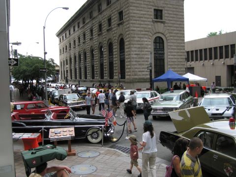 1991 Ford LTD Crown Victoria - Spectators stroll among classic police cars displayed on a city street, with vendor tents and a stone building behind.