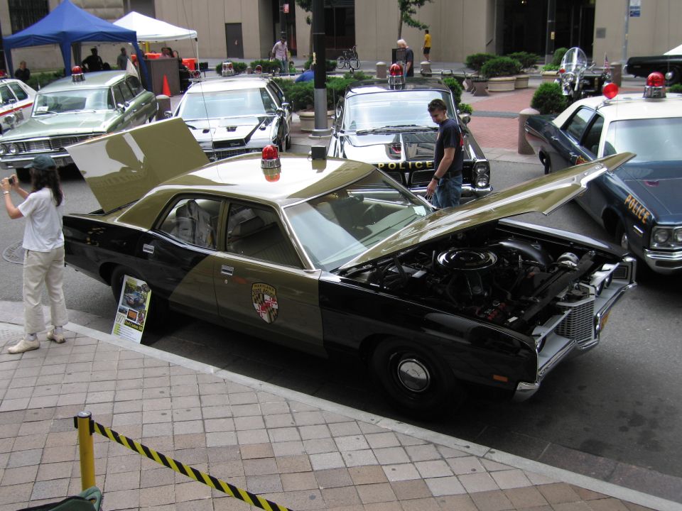 Black and tan police sedan with open hood and trunk on display at a car show