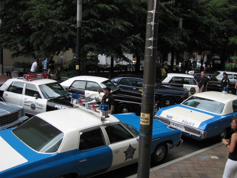 1991 Ford LTD Crown Victoria - Blue-and-white police cruisers, several marked POLICE with star emblems, are parked around a street light pole at a car show.